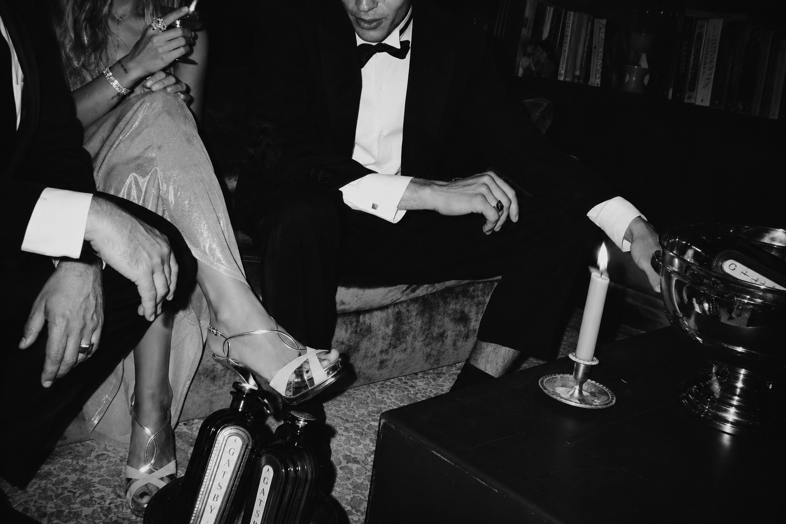 A tux and a silver-satin dress on a velvet banquette — a candle, a champagne bucket, Gatsby bottles on a low table